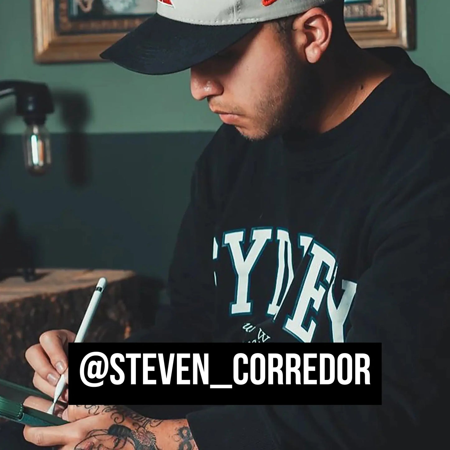 Man wearing a black 'Sydney' sweatshirt with a cap, holding a pen, in a room with a green wall.