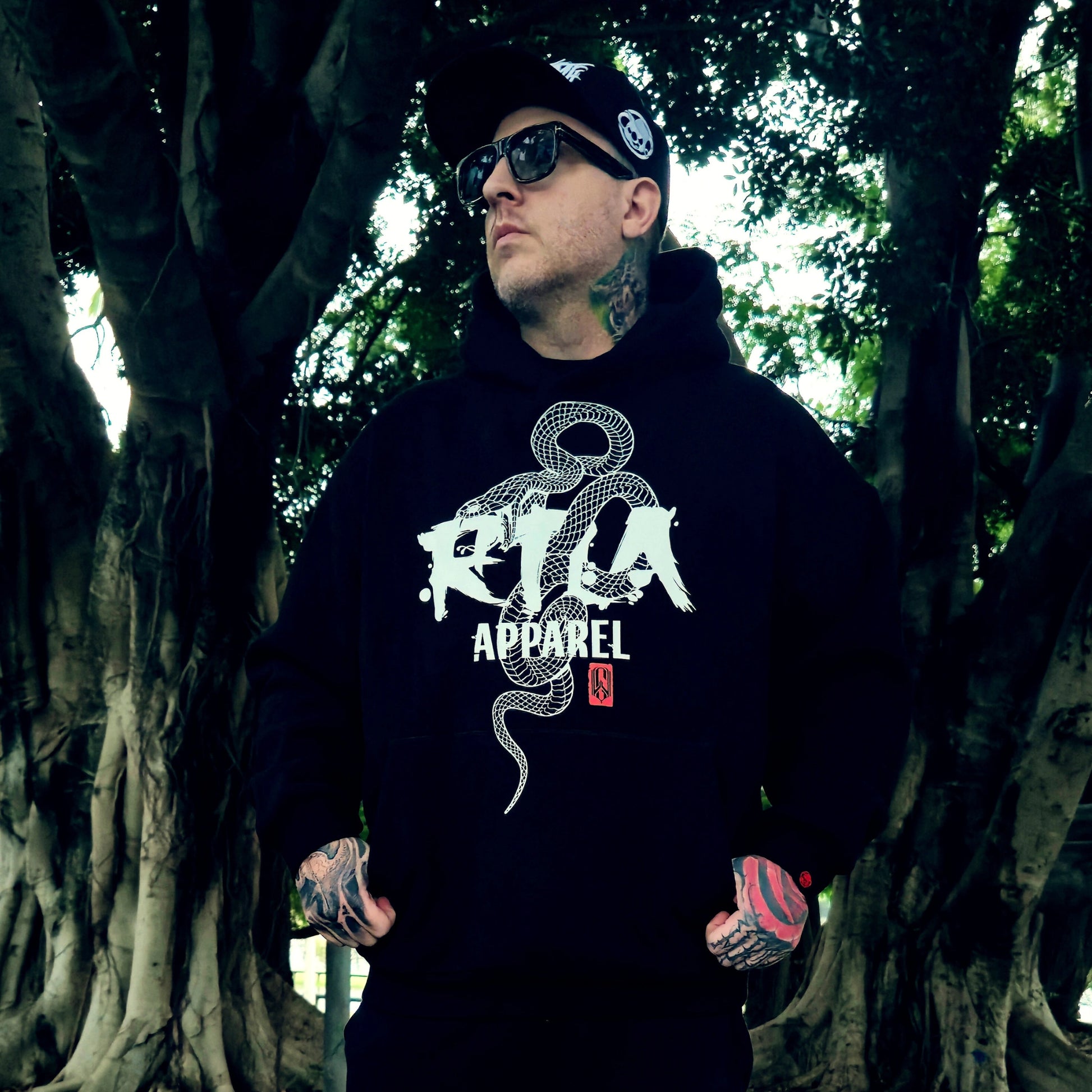 Person wearing a black hoodie with a logo and text, standing in a forest setting.