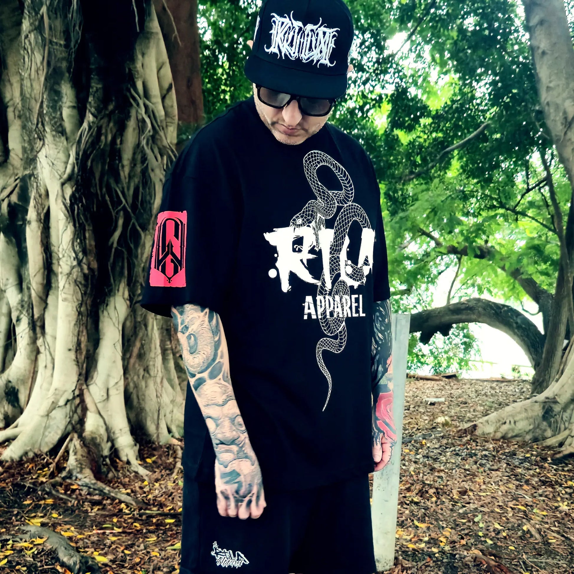 Person wearing a black t-shirt with 'Rio Apparel' logo and tattoos, standing in a forest setting.