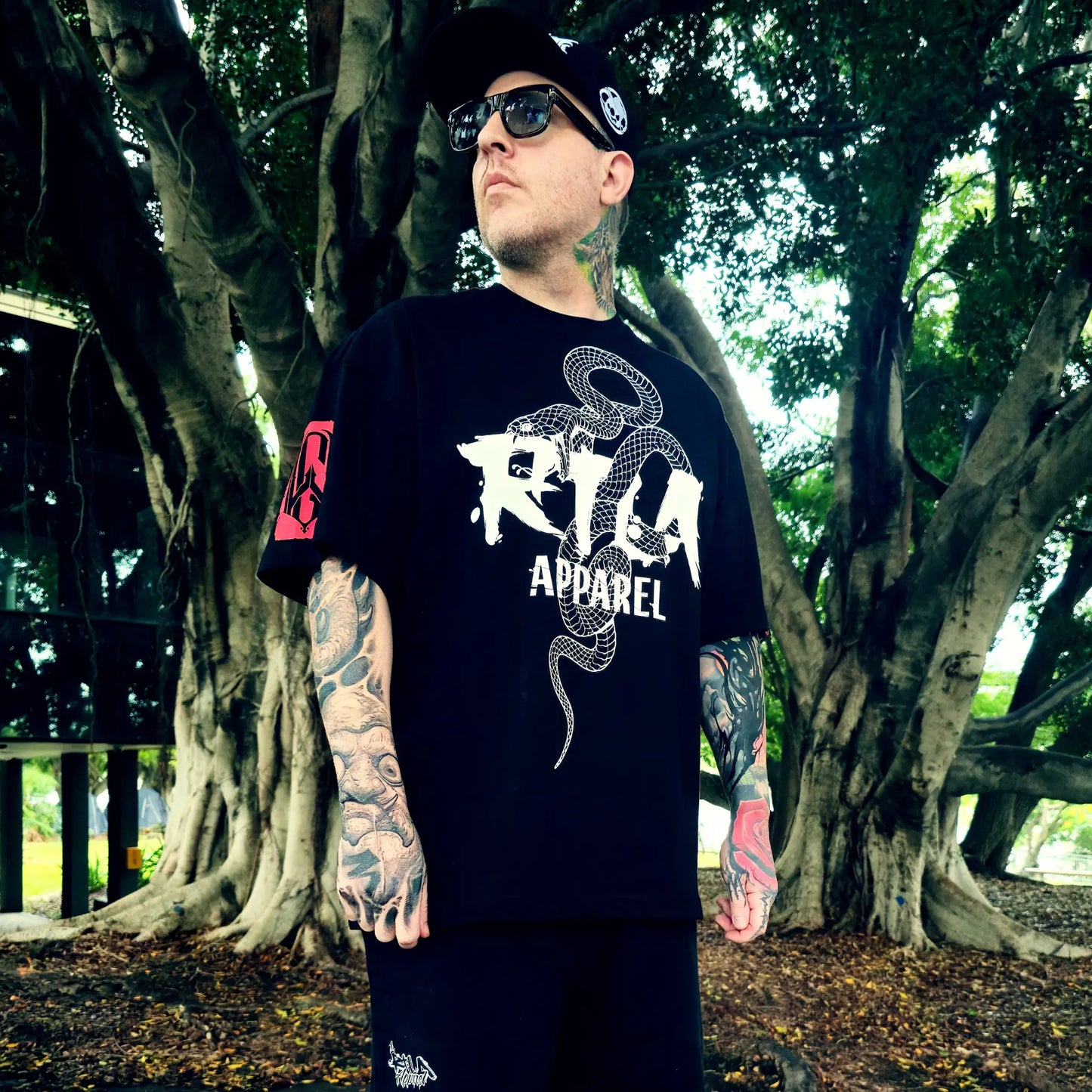 Person wearing a black t-shirt with a logo and text, standing in a forested area.